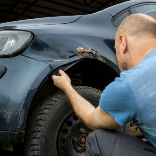 A man in a blue shirt is kneeling next to a car. He is inspecting rust damage on the wheel well.