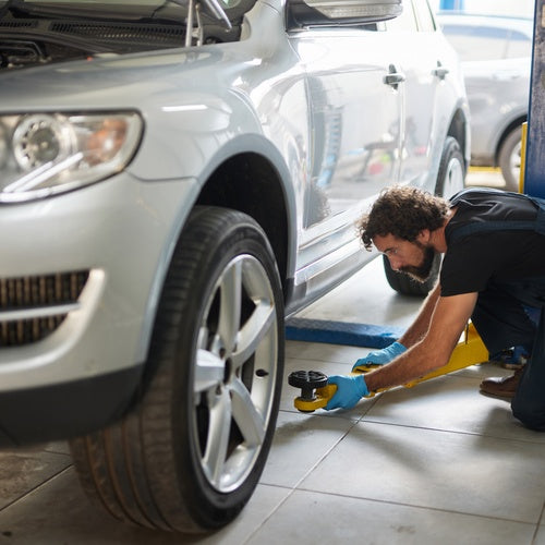 A man is putting a jack under a parked car. The car is in a garage and there are hydraulic lifts next to the vehicle.