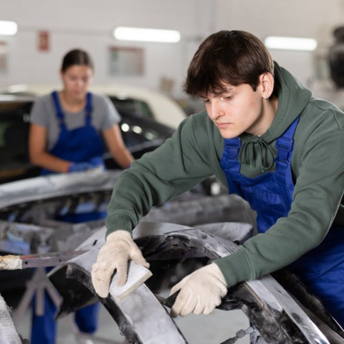 A man in blue overalls is using an abrasive tool on the frame of a car. There is a woman in the background .