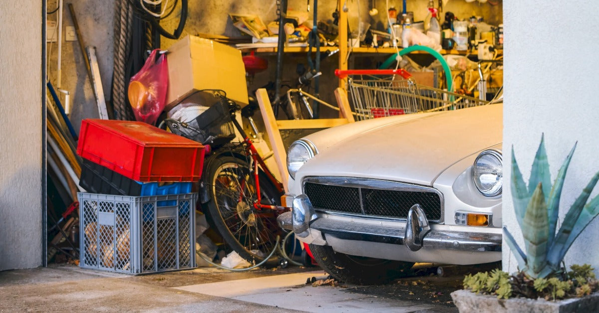 A classic car is sticking out of a garage. There are shelves filled with materials and tools.