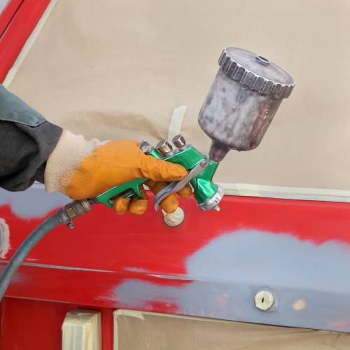 An auto body worker’s gloved hand holds an airbrush machine and paints the backside of a car bright red.