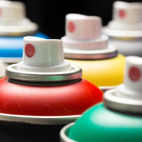 The nozzles of seven different aerosol spray paint cans stand in the air in front of a black background.