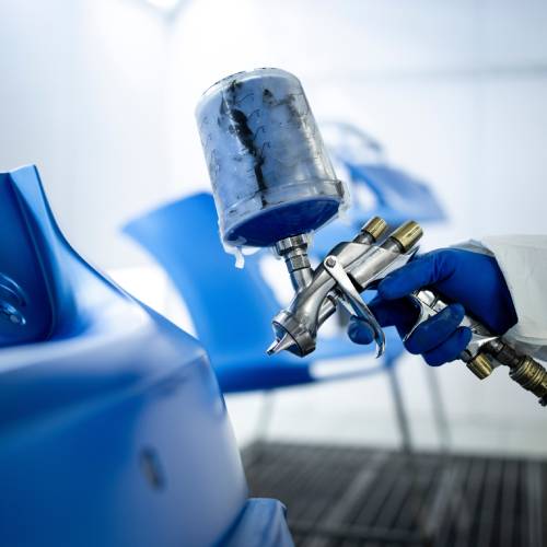 A person wearing a blue glove and white protective suit uses a spray paint gun to apply blue paint to the surface of a car.